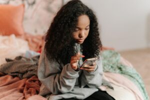 A teenage girl with curly hair using a smartphone while sitting on a bed indoors.