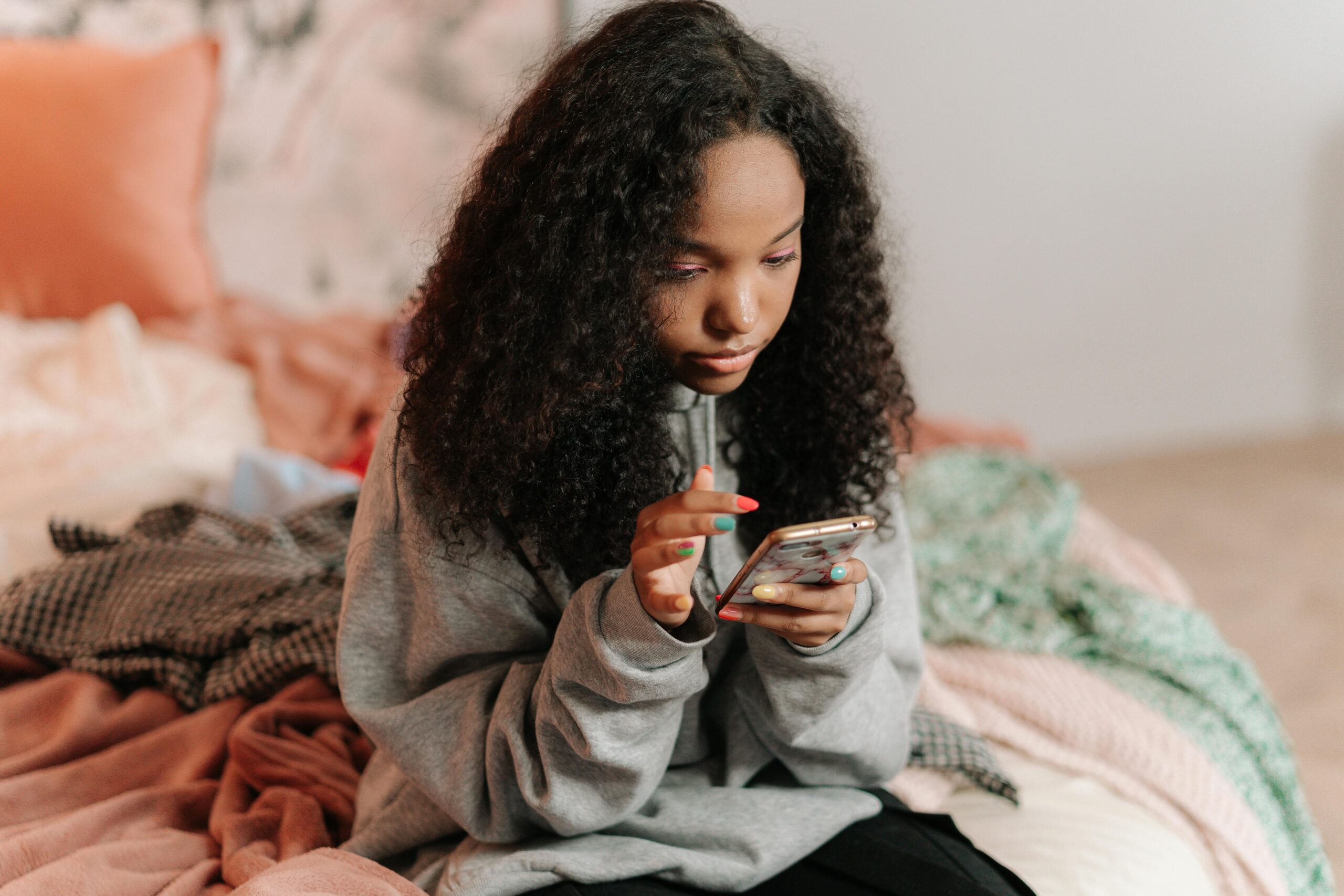 A teenage girl with curly hair using a smartphone while sitting on a bed indoors.